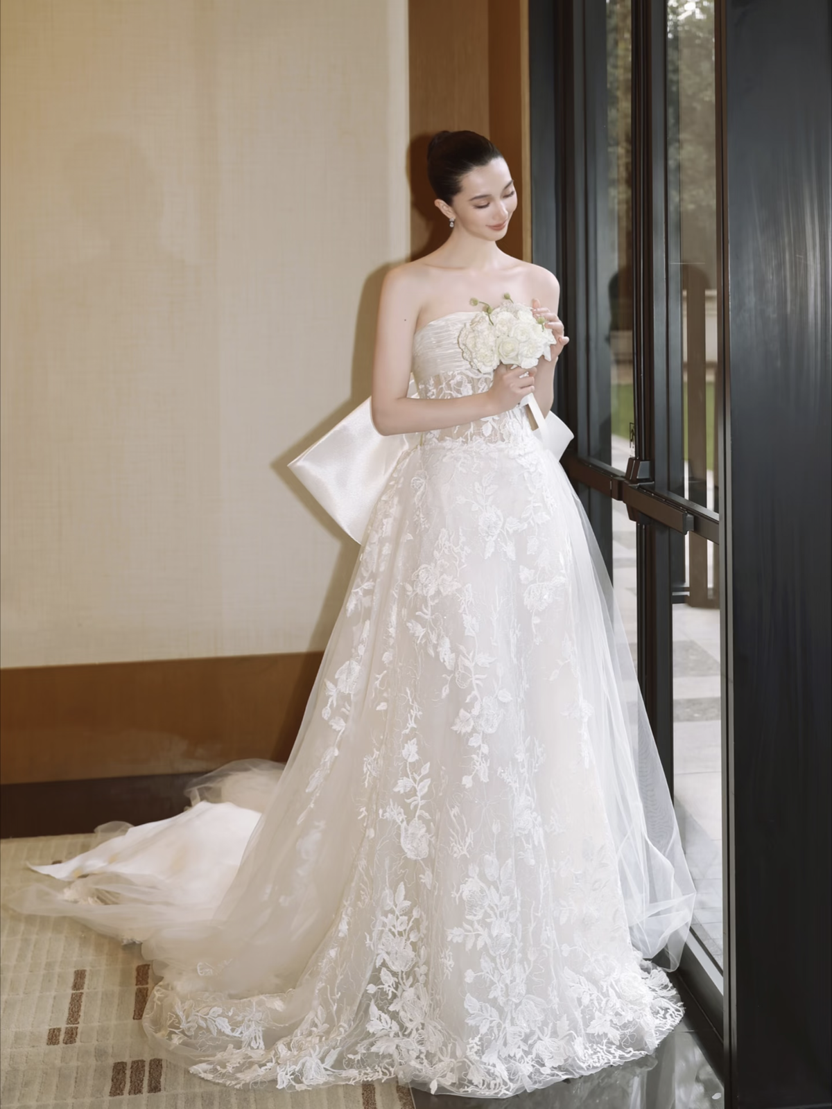 Cô dâu cầm hoa nhìn xuống váy ren trước cửa kính