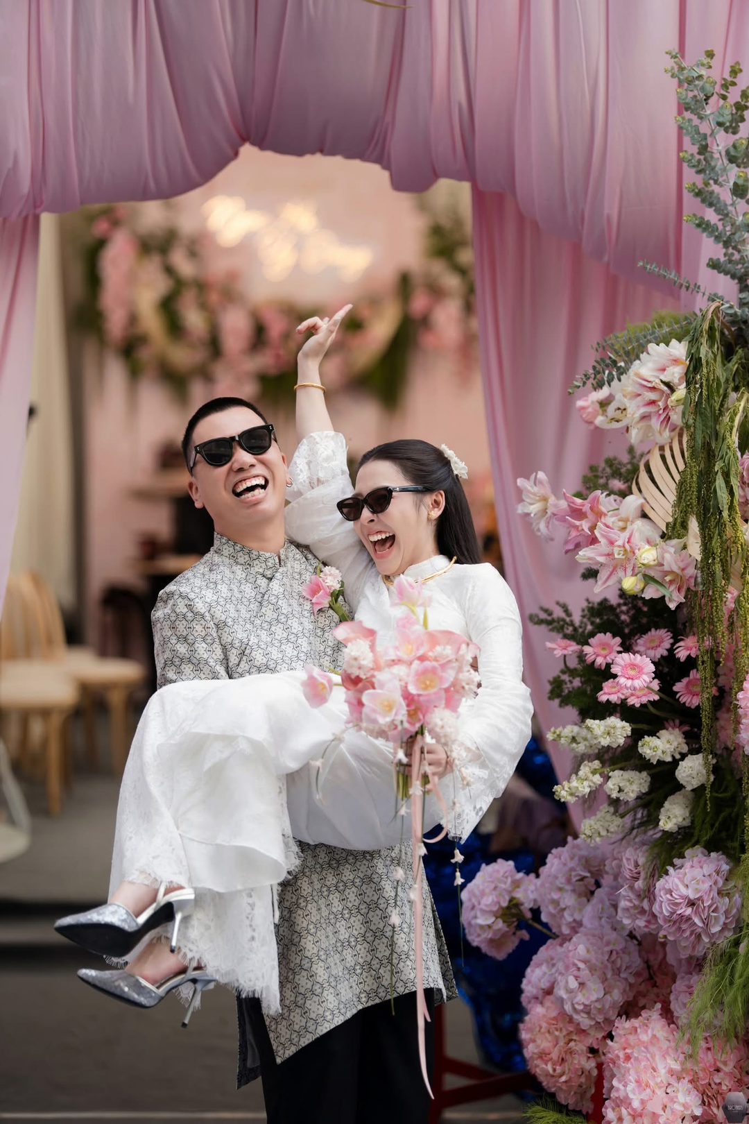 Chú rể bế cô dâu, cả hai đeo kính râm và cười bên hoa hồng